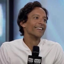 An image of Danny Pudi smiling and holding a microphone during an interview. Danny has brown skin and wavy black hair. He is wearing a white shirt. The background is light blue.